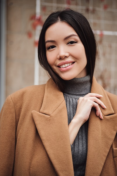 Portrait of a gorgeous Japanese girl in a brown coat happily looking in a camera outdoors