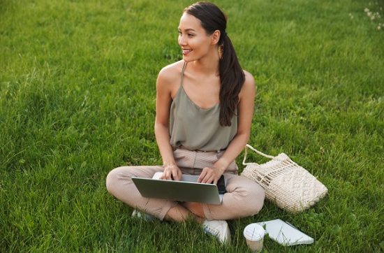 Pretty smiling Japanese woman sitting on grass with a laptop on her knees