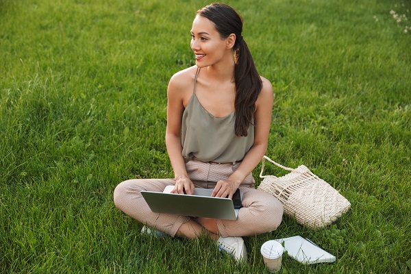 Pretty smiling Japanese woman sitting on grass with a laptop on her knees